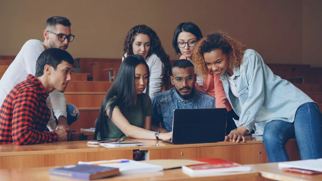 A group of young students leaning in around a laptop, working through a problem together