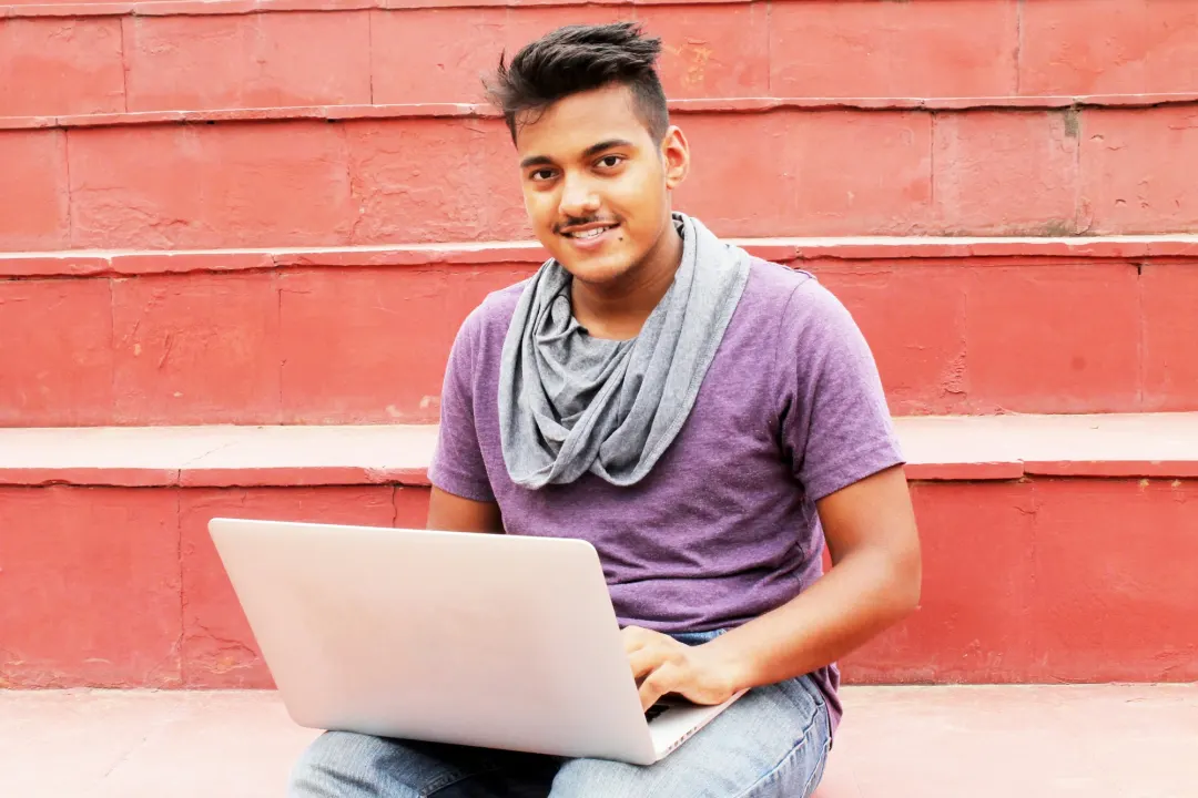 A young developer smiling while working on a laptop, sitting on campus steps
